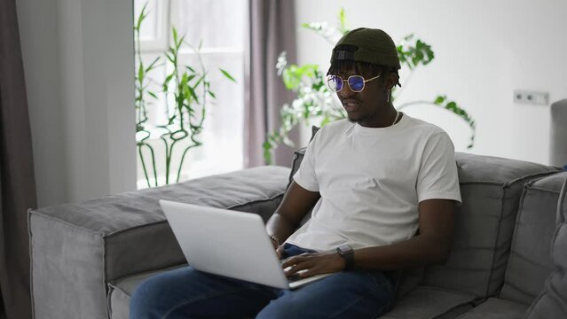 Attractive african American man in glasses sitting on the sofa using laptop at home