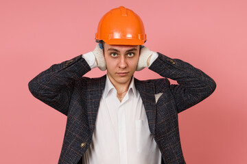 a male engineer in a suit and a protective helmet does not hear anything, covering his ears with his hands on a colored background