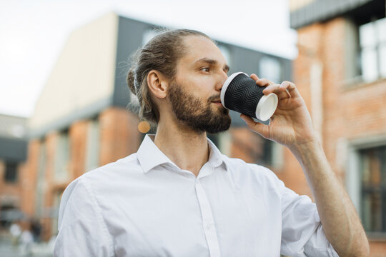 Closeup confident business man drinking coffee to go in city. Portrait of beautiful handsome man worker looking away outdoors. Serious businessman waiting client on street.