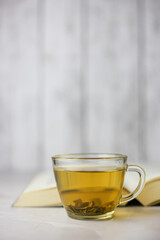 Brewed herbal green tea in a transparent cup in front of an open book on a light background.
