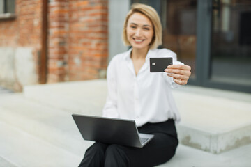 Fototapeta premium Focus on bank credit card. Business payment of bills concept. Smiling young woman, using notebook and holding credit card for making payments online. Female freelancer receive payment online.