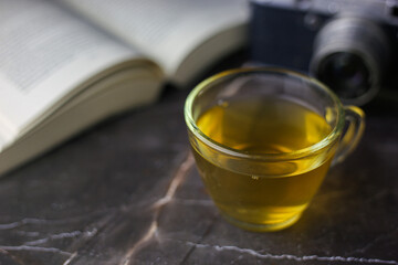 Brewed herbal green tea in transparent cup near open book and vintage camera on dark background