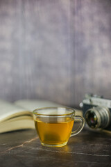 Brewed herbal green tea in transparent cup near open book and vintage camera on dark background