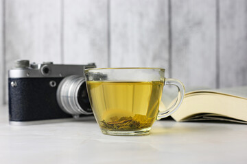 Brewed herbal green tea in a transparent cup next to an open book and a vintage camera on a light background