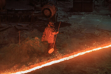 Worker passing along red hot slag stream at steel works.
