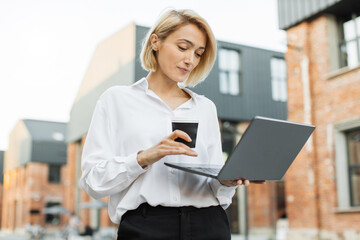Portrait of cheerful caucasian woman having cup of take away fresh beverage talking to partner having online video chat using laptop walking on city street looking at screen.