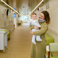 Mother woman with toddler baby boy in her arms visit in the hall clinic. Mom with a child in the...