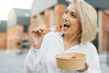 Happy positive business woman eating healthy salad on a break standing over city street background. Female dieting nutrition concept. Attractive smiling girl enjoying veggie meal.