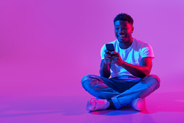 African American guy sitting cross legged with smartphone, surfing social media, chatting to friend...