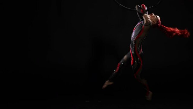 Flexible slim young woman in stage costume spinning in slow motion holding on air hoop with hands. Wide shot portrait of skilled talented Caucasian aerial gymnast performing modern choreography dance