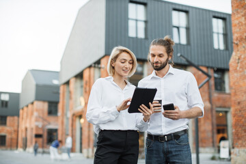 Charming woman with digital tablet and attractive man with smartphone and cuo of take away coffee standing together near office building. Two colleagues using modern gadgets for work outdoors.