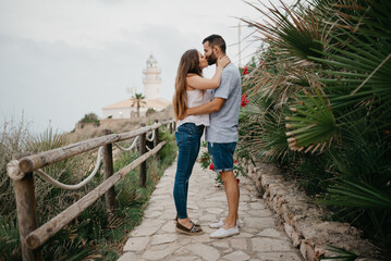 A Hispanic man and his Latina girlfriend are kissing each other on the path from a lighthouse between palms in Spain. A couple of tourists are enjoying each other on a date at sunset in Valencia.
