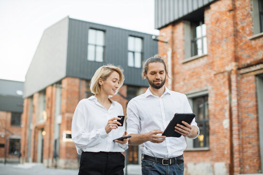 Two Caucasian Business People Conducting Financial Transactions Online Outdoors. Blond Lady Using Credit Card And Smartphone For Cashless Payment, Her Male Colleague Holds Tablet.