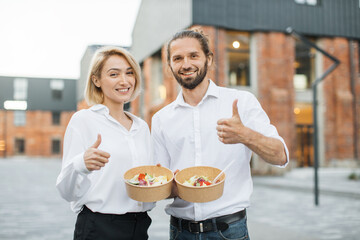 Young cheerful couple, colleague or business partners in white shirts having break and eating healthy meal walking at the city street showing thumb up. Organic healthy nutrition concept.