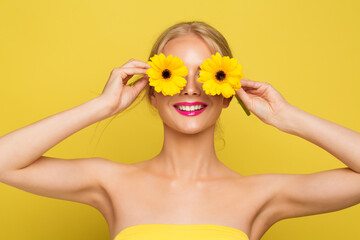 Woman covering Eyes with Yellow Flowers. Happy Smiling Model with Gerbera Floral Sun Glasses over isolated Orange background. Cheerful Summer Fashion Girl