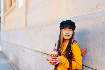 young asian woman with backpack using her mobile phone and drinking a chocolate shake in the street. chinese girl doing tourism in europe