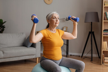Fototapeta na wymiar Stay home, do sports. Fit senior woman doing exercises on fitness ball and using dumbbells, working out at home
