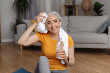 Happy senior woman with bottle of water wiping sweat with towel after home workout, smiling at...