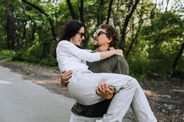 Obraz premium A bearded long-haired stylish man holds his beloved hippie brunette woman in sunglasses in his arms and circles her in nature in a park on the road. Portrait, photo of happy and smiling newlyweds.