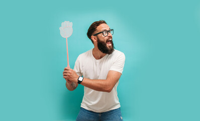 Young caucasian charismatic man holding a fly swatter wanting to kill annoying mosquito or a fly. - obrazy, fototapety, plakaty