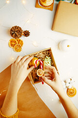 Woman's hand puts golden Christmas ball and cinnamon with candy cane in gift box. Top view of white table with festive decorations, wrapping paper, fir cones, dried oranges. New Year atmosphere.