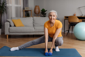 Happy senior woman warming up, doing legs stretching exercises while working out at home on fitness...