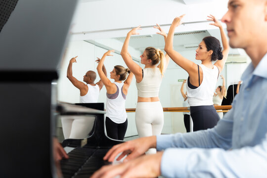 Group of multiethnic dancers training with one hand on barre under live music in .bright fitness room