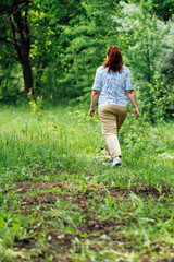 Back view of young buxom woman with long curly red hair in ponytail, wearing blue blouse with floral pattern, beige trousers, walking on path in park forest among green trees. Summer, nature, relax.
