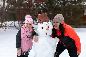 Portrait of little girl and woman with built snowman with snow smiling with teeth in winter while having walk in park with trees in background. Parents spending time with children.