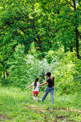 Back view of happy family walking in park forest around green trees, having fun. Little cheerful daughter holding hand of middle-aged bearded man father. Love, summer activities, travelling. Vertical.