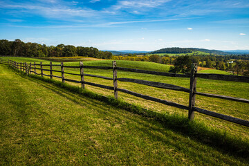 Fototapeta na wymiar Meadow with wooden fence, green grass, hilly landscape and blue sky with white clouds on a beautiful summer day in the Sky Meadows State Park, Virginia, USA.
