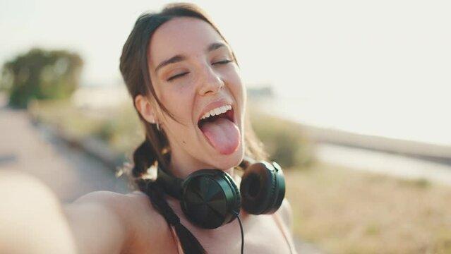 Cute young athletic woman with braided pigtail wearing beige sports top with headphones stands on the embankment. Beautiful girl crosses her hand and smiles at the camera