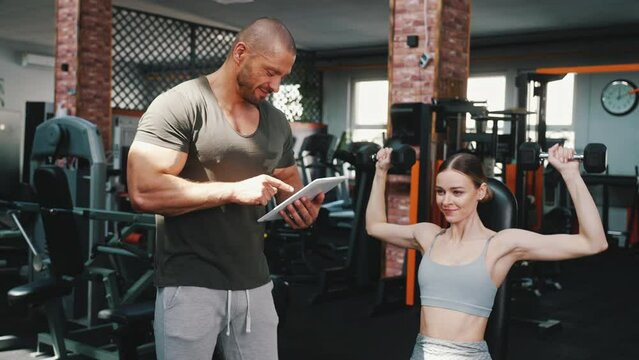 Fit beautiful caucasian young adult woman in gray sports bra lifting dumbbell while sitting on bench. Muscular personal trainer conducts a fitness test. High quality 4k footage