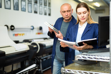 Female publishing facility worker reading operation manual for large printer. Her male co-worker standing beside. - obrazy, fototapety, plakaty