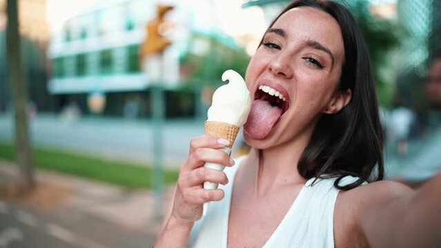 Close-up of beautiful woman with freckles and dark loose hair wearing white top is testing ice cream and taking selfie. Cute girl enjoys ice cream on modern city background