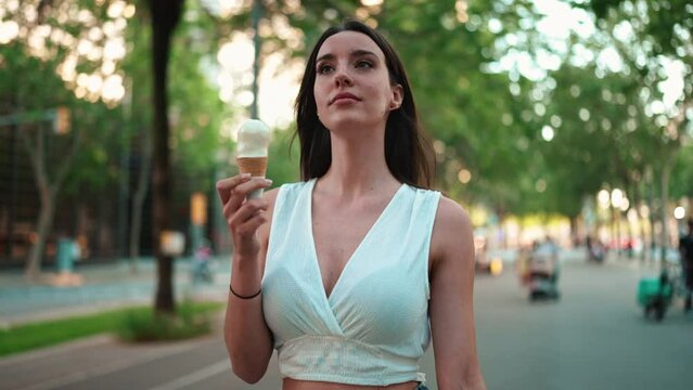 Beautiful Woman With Freckles And Dark Loose Hair Wearing White Top Is Walking Down The Street And Testing Ice Cream. Cute Girl Enjoys Ice Cream On Modern City Background 