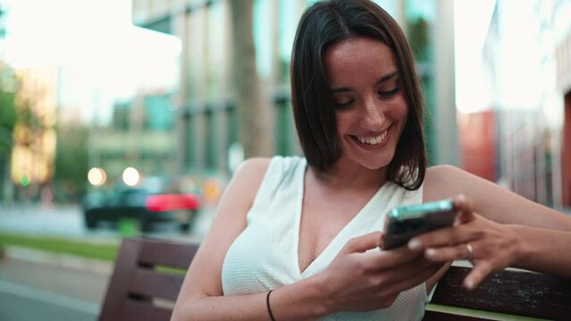 Beautiful woman with freckles and dark loose hair wearing white top sits on bench and watching messages, photos and videos on mobile phone. Camera is getting closer