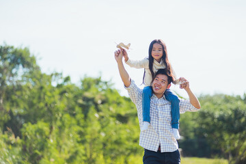 Happy Asian young family father and carrying an excited girl on shoulders having fun and enjoying outdoor lifestyle together playing aircraft toy on sunny summer day, Father's day concept