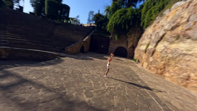 Drone shot of a girl in a white tight suit doing leg-split in the amphitheater