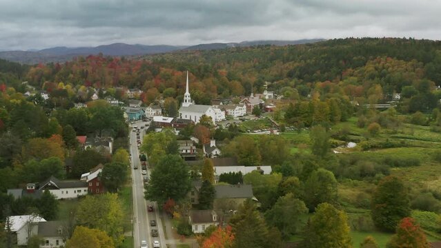 Main street to Church building in small town with fall foliage forest landscape background on rainy day. East Coast Vermont USA. Cinematic 4K aerial of white wooden old historic Stowe community church