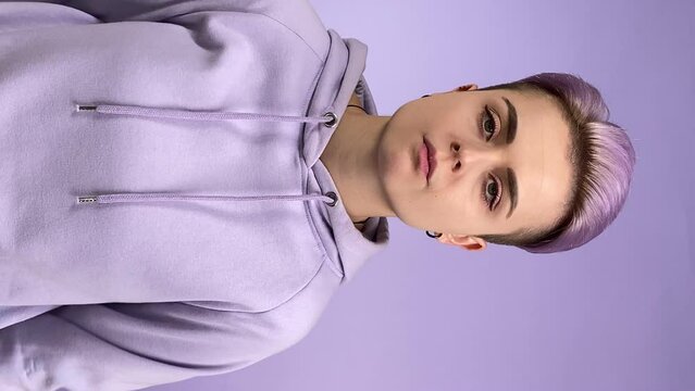 Trendy young adult woman generation Z having indifferent look, chewing bubblegum, folding arms crossed on purple background indoors. Frigid female person with short hair looking at camera dismissively