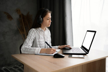 Young female student in earphones sit at desk at home using laptop screen studying making notes, concentrated Hispanic girl in headphones watch webinar on computer, distant education