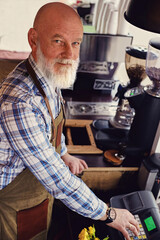 Shot of old man barista dressed in plaid shirt and apron looking at camera inside coffee shop.