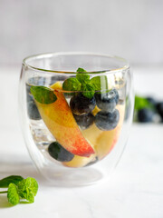 Blueberry and peach infused water, cocktail, lemonade or tea in glass. Summer iced cold drink with blueberry, ice, peach and mint on grey marble background, copy space, vertical, close up, side view