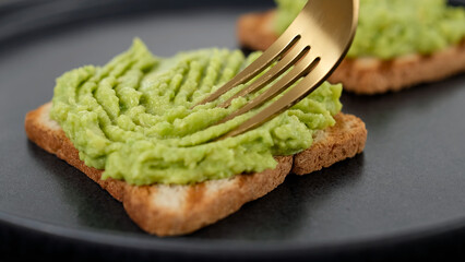 Preparing avocado toast. Hands with fork spreading mashed avocado on toasted whole  bread. Vegan food concept