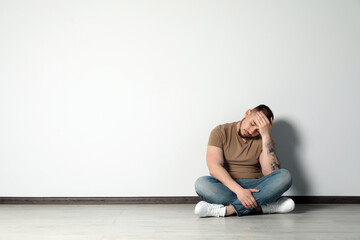 Sad young man sitting on floor near white wall indoors, space for text
