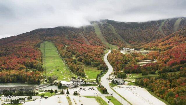 Mountain resort with scenic fall foliage forest landscape background on cloudy autumn day. Cinematic 4K aerial white Killington ski resort area with hotels and golf course on sunny colorful fall day