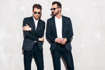 Portrait of two handsome confident stylish hipster lambersexual models. Sexy modern men dressed in black same elegant suit. Fashion male posing in studio near grey wall