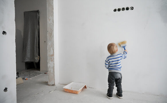 Back view of little boy painting white wall in empty room under renovation. Cute kid using paint brush while renovating apartment. Concept of home renovation, restoration and childhood.