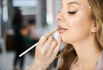 Makeup artist doing professional makeup for client with braces on teeth. Close up of female hand in sterile glove applying lipstick on woman lips with cosmetic lip brush.